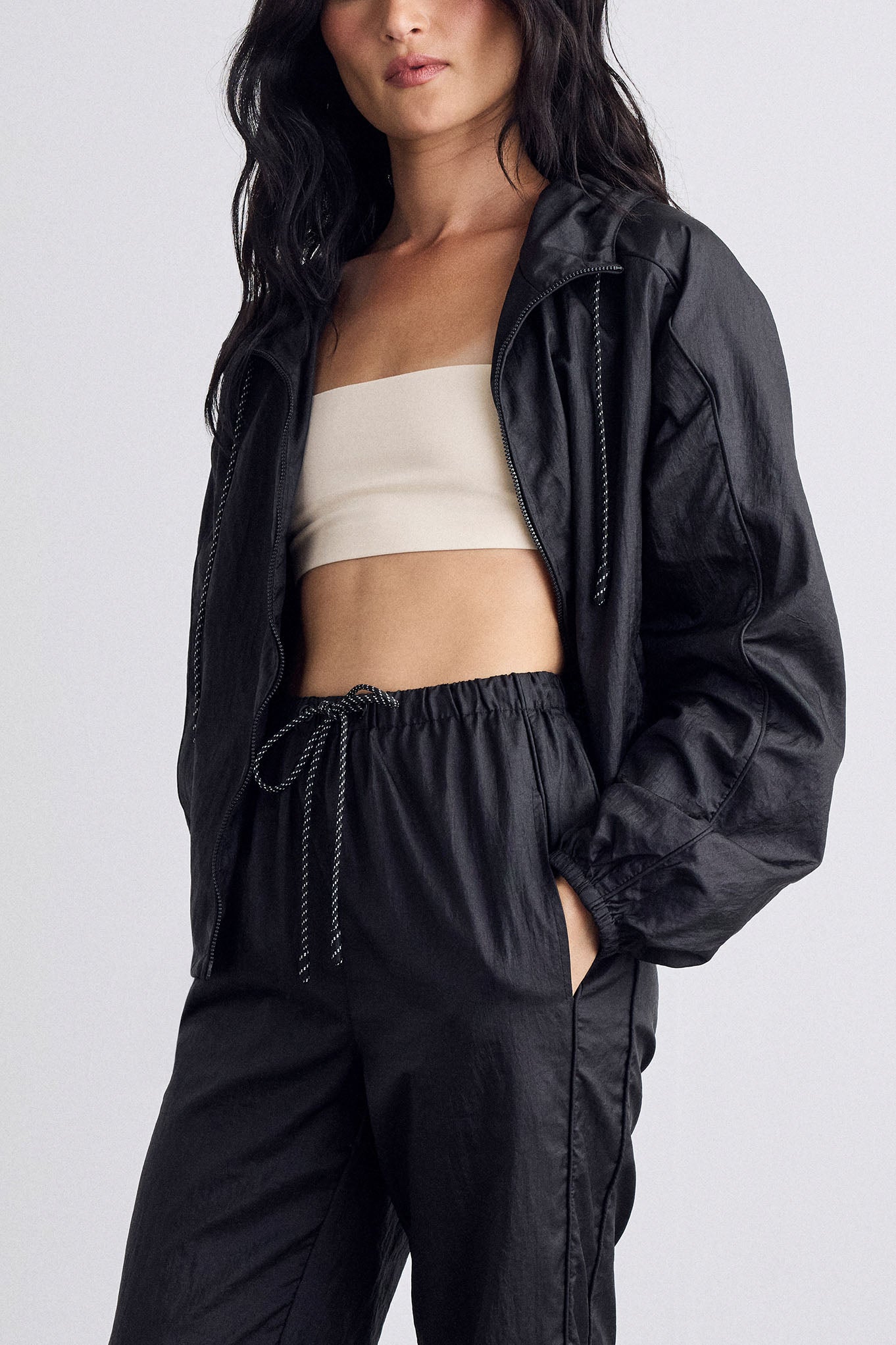 A model wearing a black lightweight zip-up hooded jacket and matching black drawstring sweatpants, paired with a  cream sports top, against a light gray studio background.