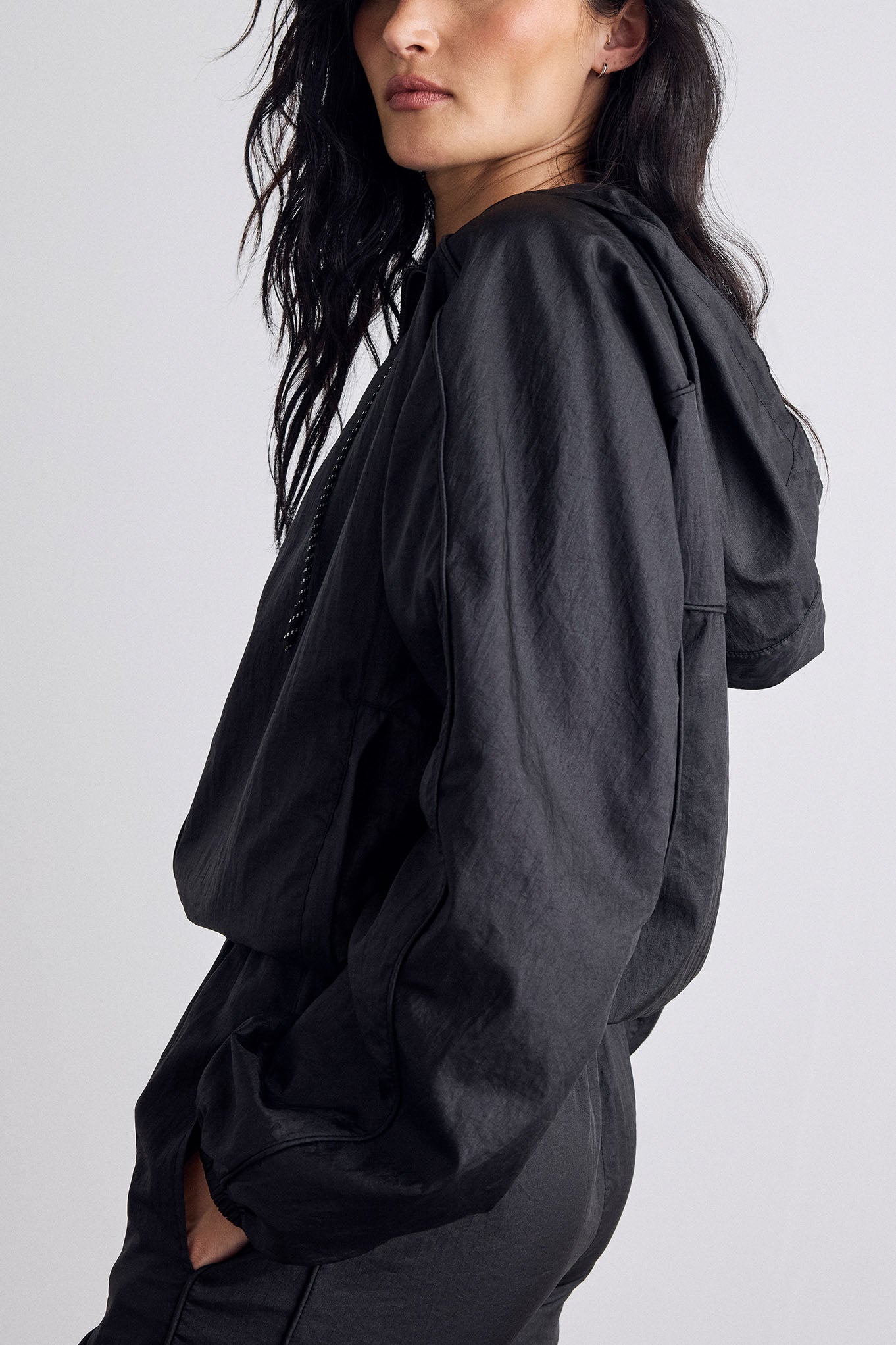 A woman wearing a black hooded jacket, shown from a side-back angle against a light gray studio background.