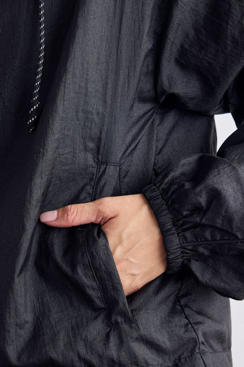 Close-up of a person wearing a black lightweight jacket, with one hand tucked into a front pocket. The fabric is slightly sheer and crinkled, with elasticated cuffs on the sleeves.