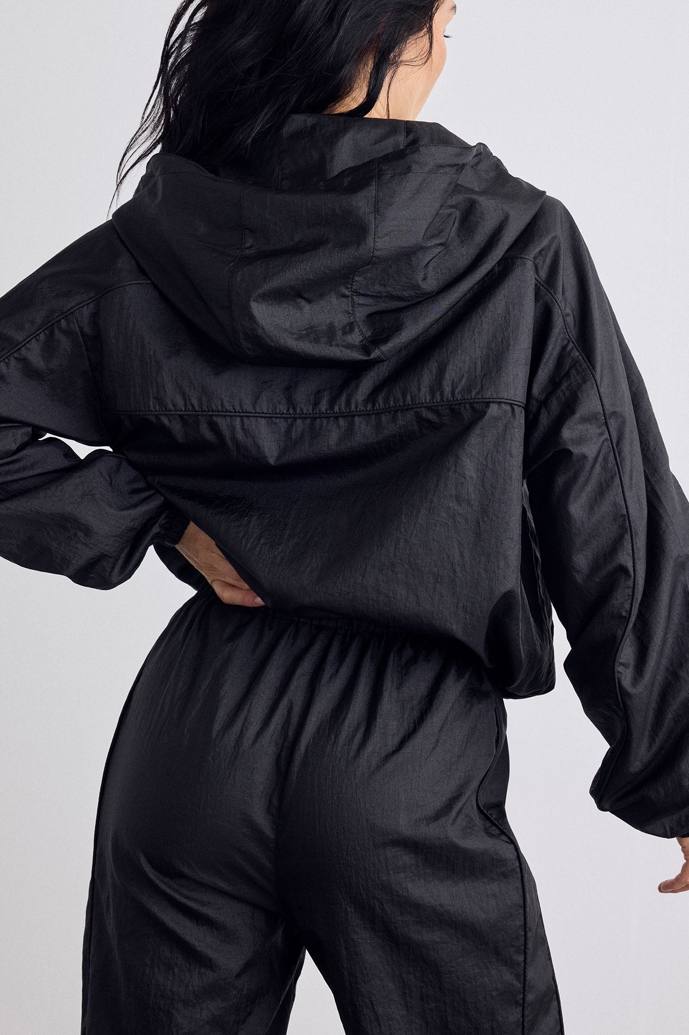 A person shown from the back wearing a black hooded jacket and matching black pants against a light gray studio background.