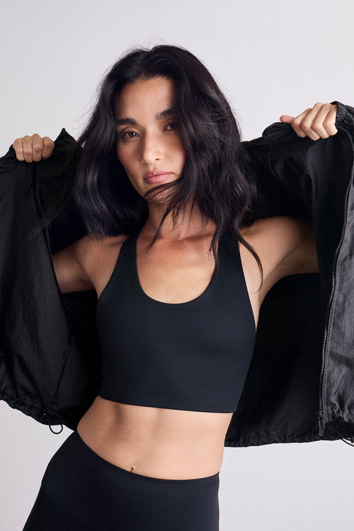 A woman modeling a black athletic sports bra and black leggings, holding open a black puffer jacket against a light gray studio background.