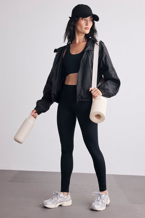 A woman modeling an athletic outfit consisting of a black sports bra, black leggings, and a black lightweight jacket, paired with a black cap and carrying a rolled cream yoga mat and a cream water bottle against a light gray studio background.