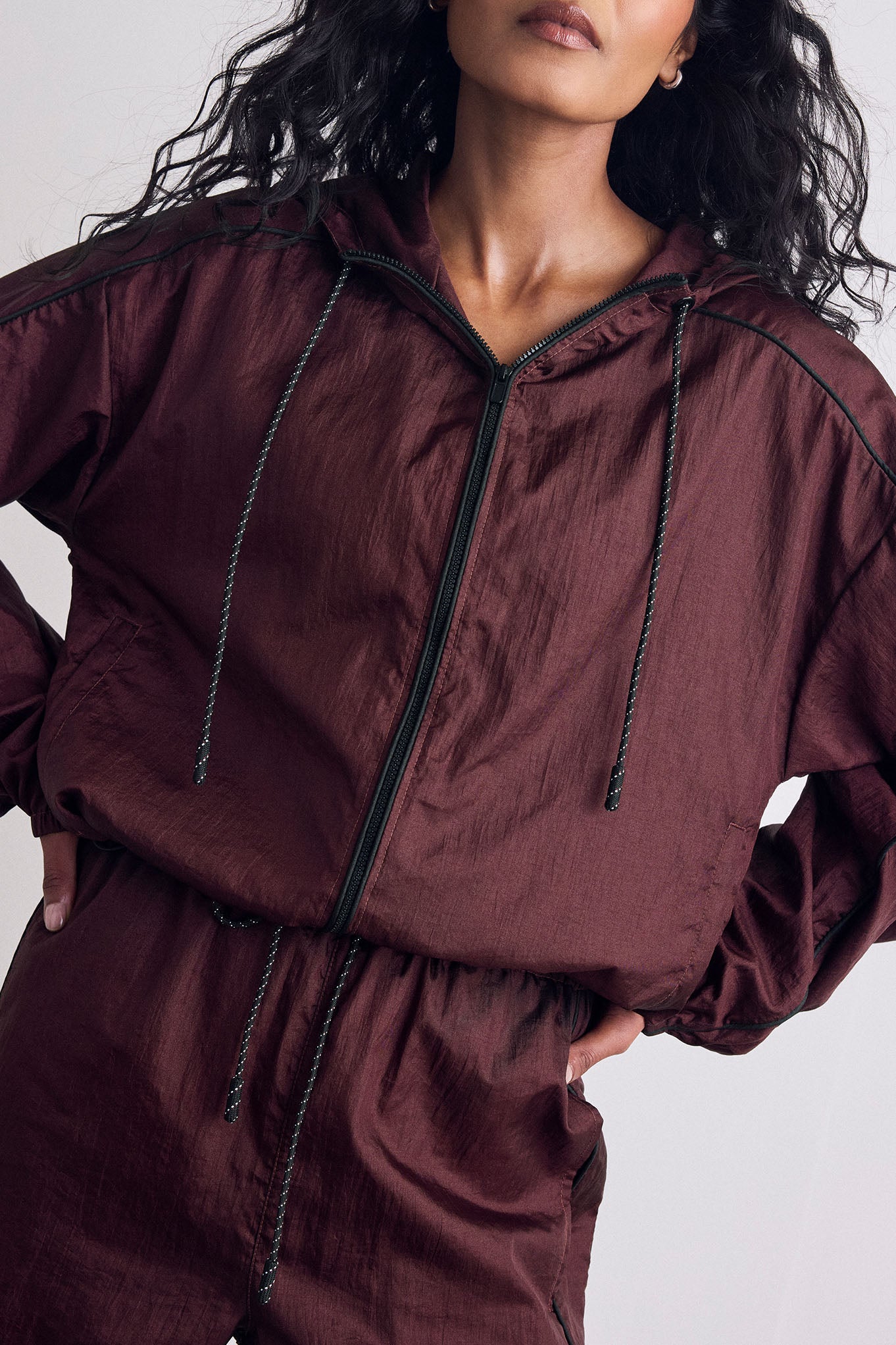 A close-up of a model wearing a lightweight, crinkled burgundy hooded windbreaker jacket with black drawstrings and a black front zipper, set against a plain light gray background.