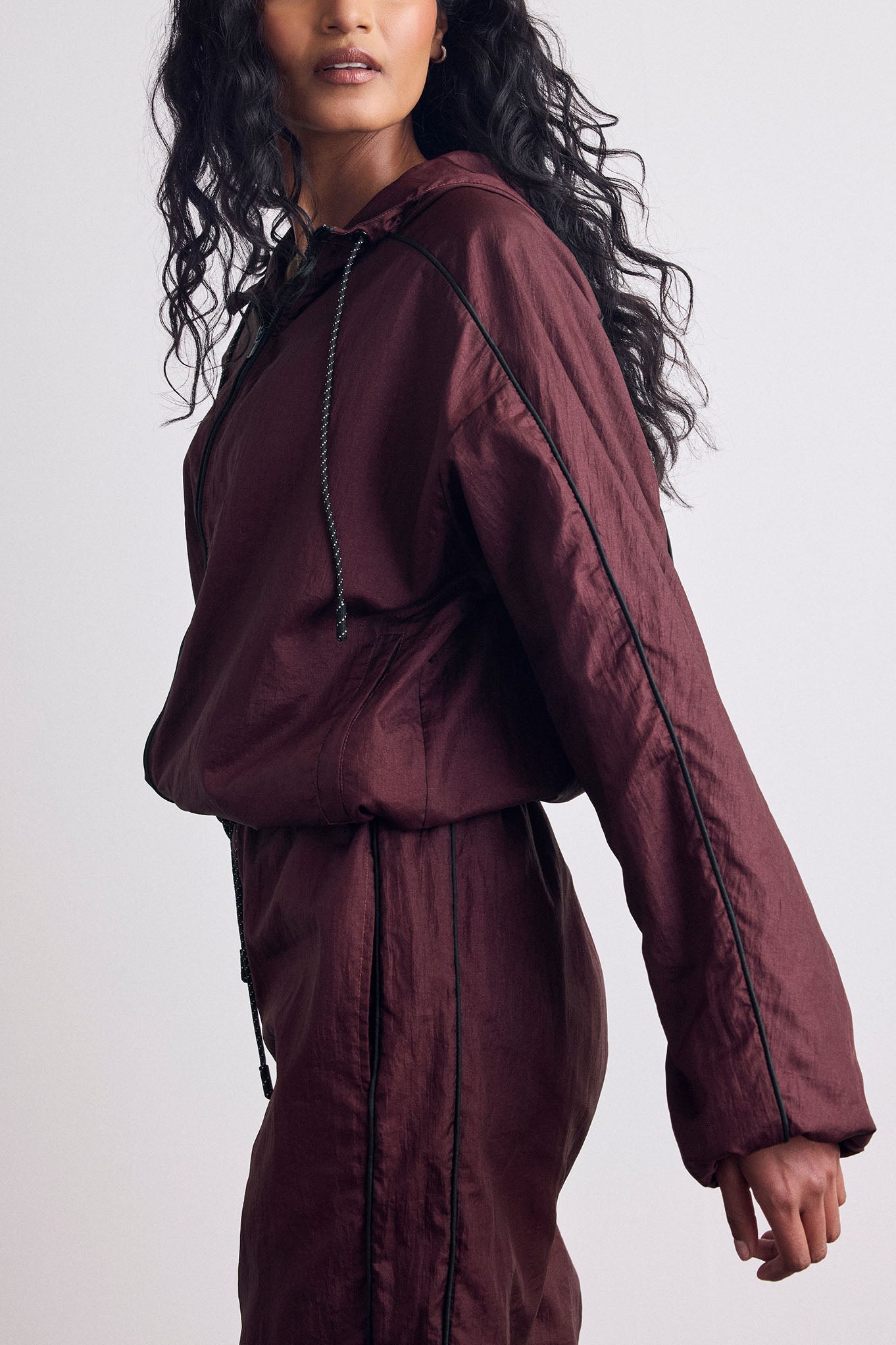 A model wearing a matching burgundy hooded windbreaker jacket and pants set with black piping and drawstrings, shown from the side against a light gray studio background.