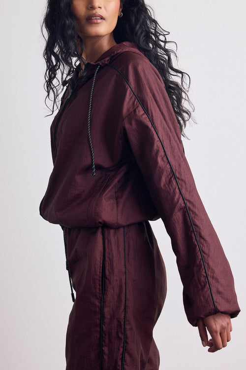 A model wearing a matching burgundy hooded windbreaker jacket and pants set with black piping and drawstrings, shown from the side against a light gray studio background.