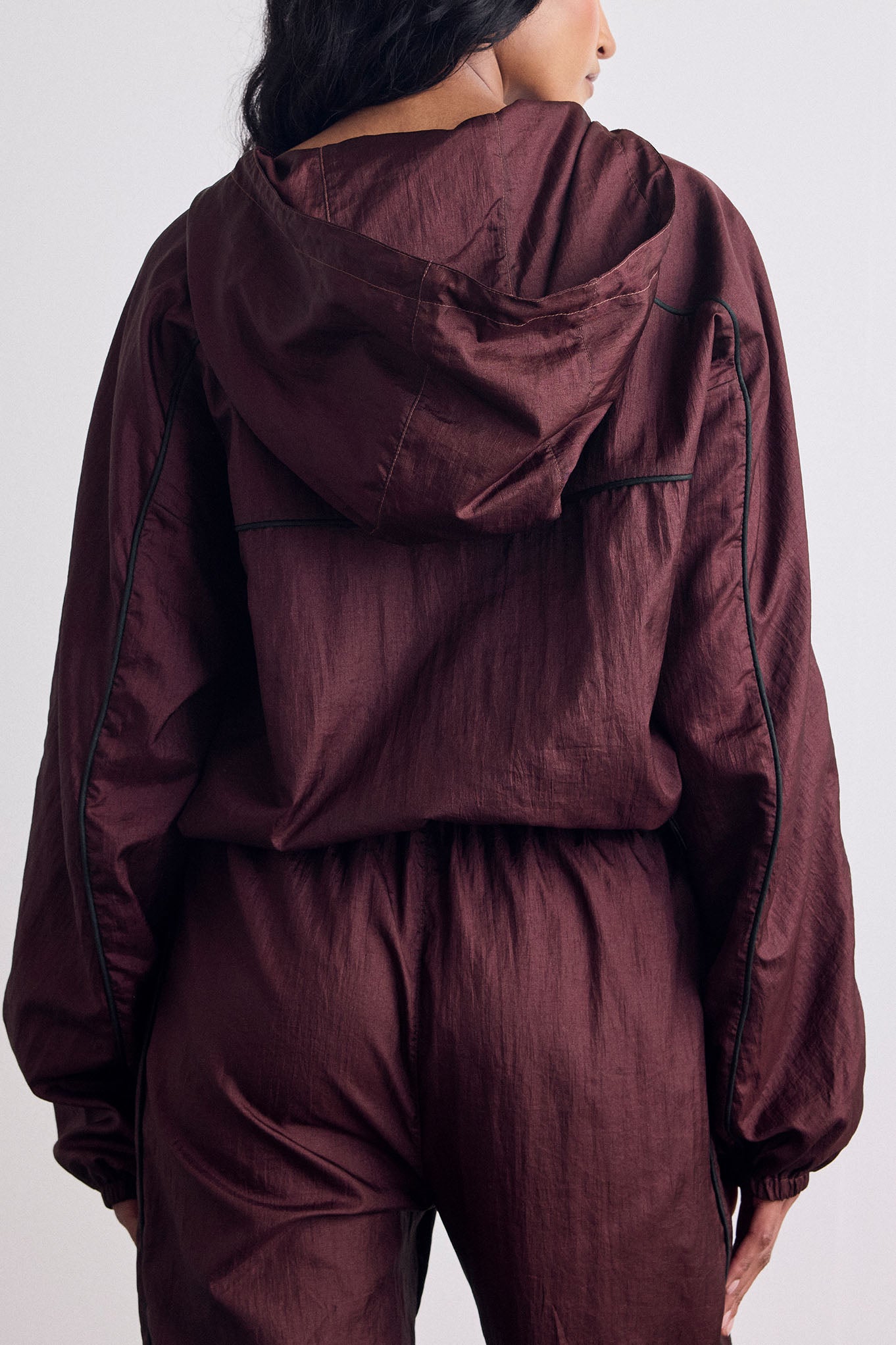 A person shown from the back wearing a matching burgundy hooded jacket and jogger-style pants with thin dark piping along the seams, against a plain light gray studio background.