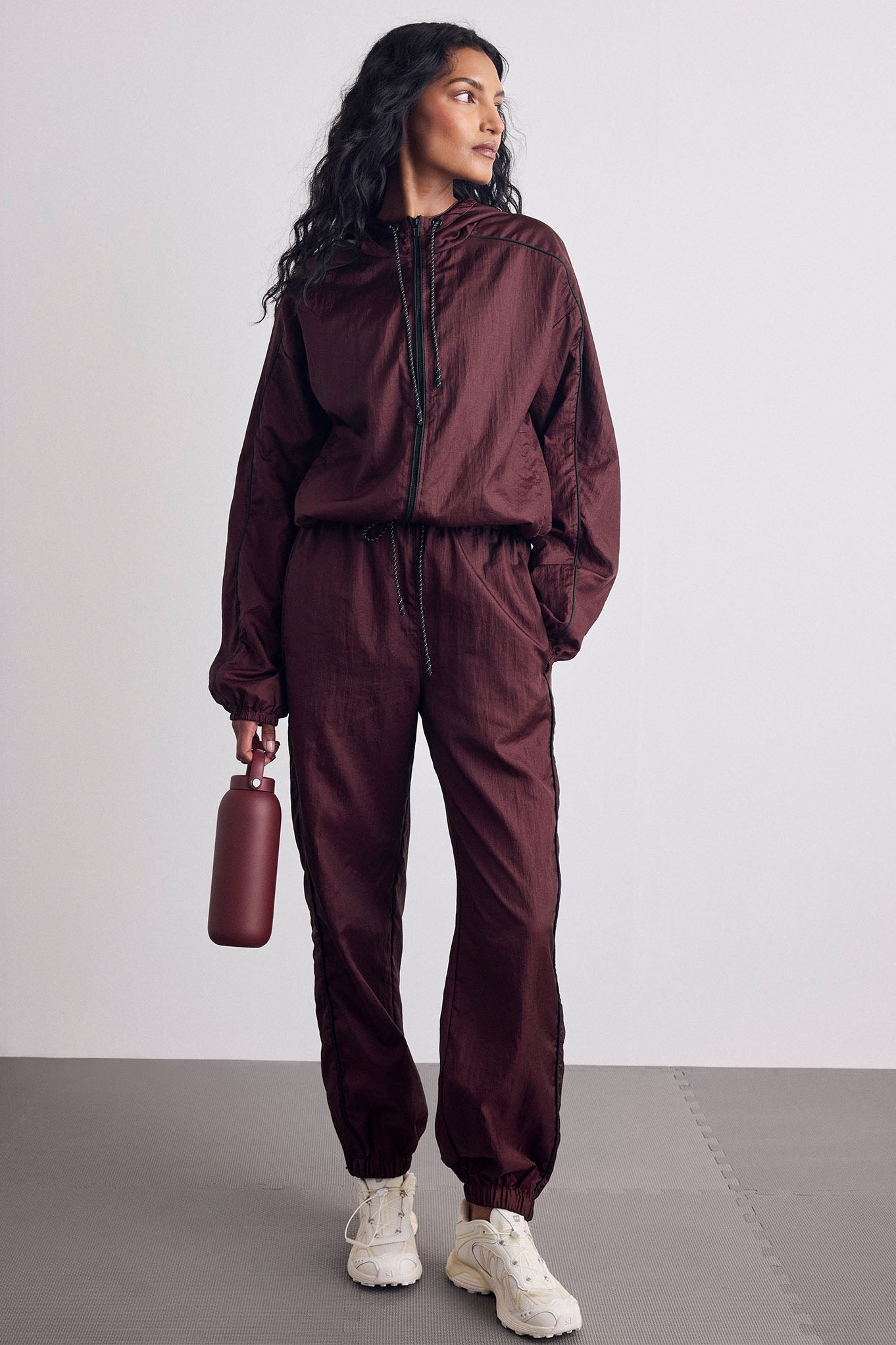 A woman modeling a matching burgundy satin tracksuit consisting of a zip-up jacket and jogger pants, paired with white sneakers and holding a matching burgundy bottle against a light gray studio background.