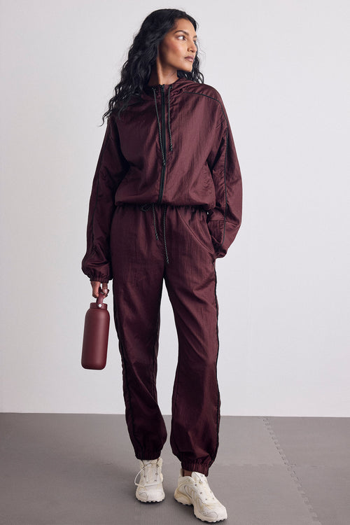 A woman modeling a matching burgundy satin tracksuit consisting of a zip-up jacket and jogger pants, paired with white sneakers and holding a matching burgundy bottle against a light gray studio background.