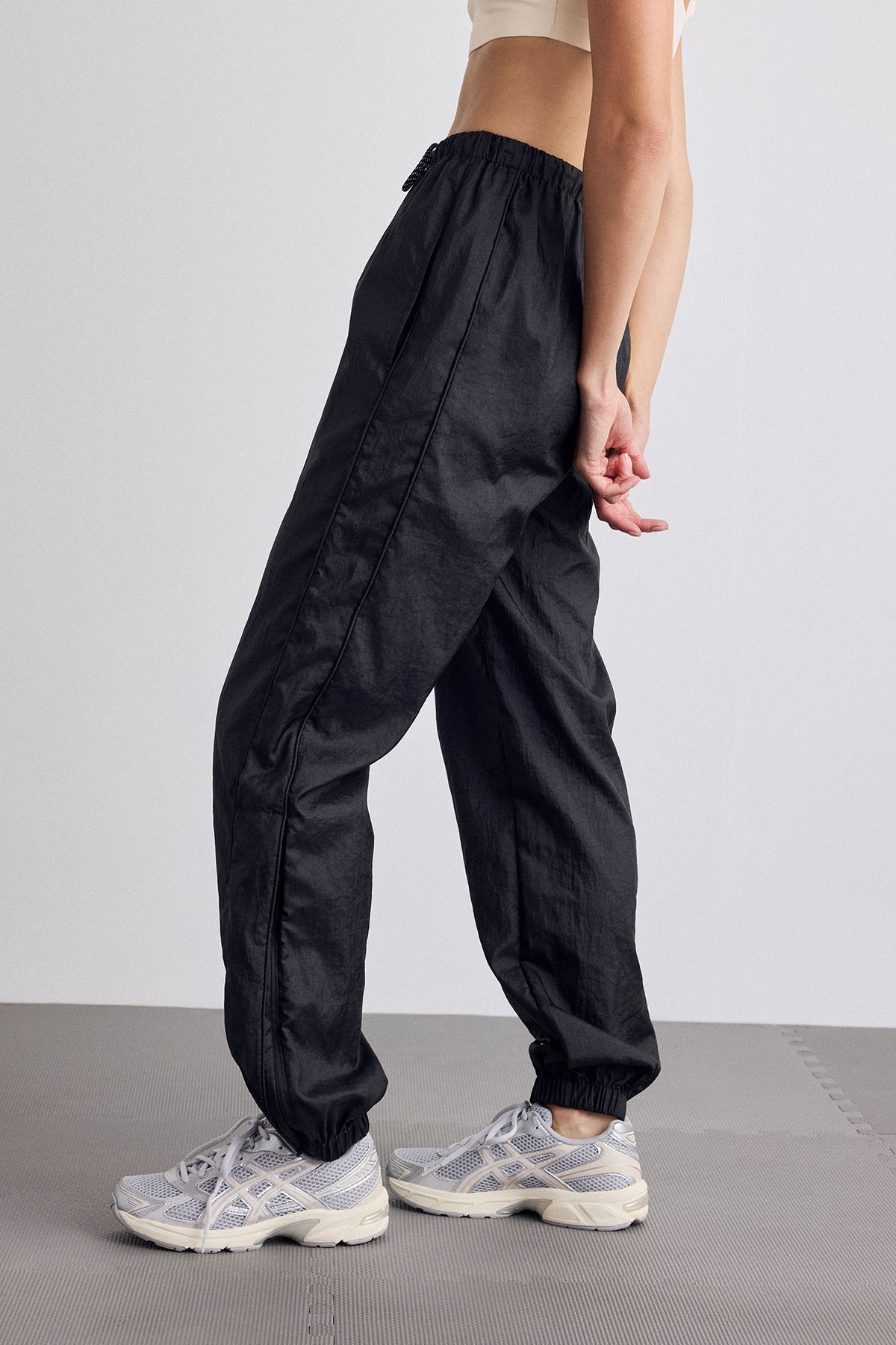 A person wearing loose black jogger-style pants with an elastic waist and cuffs, paired with light gray running shoes, shown from the waist down against a plain light gray studio background.