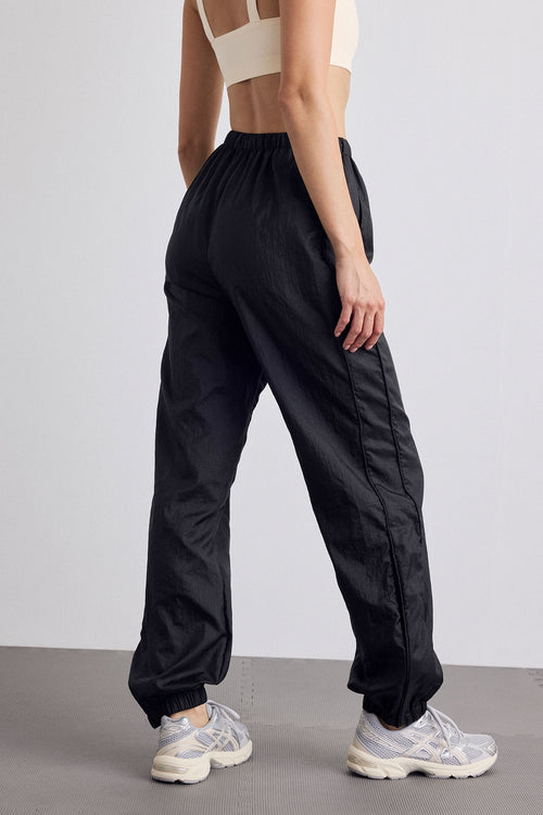 A model from the back wearing black wide-leg jogger pants with elastic cuffs, paired with a cream sports bra and white sneakers, set against a minimalist studio background.