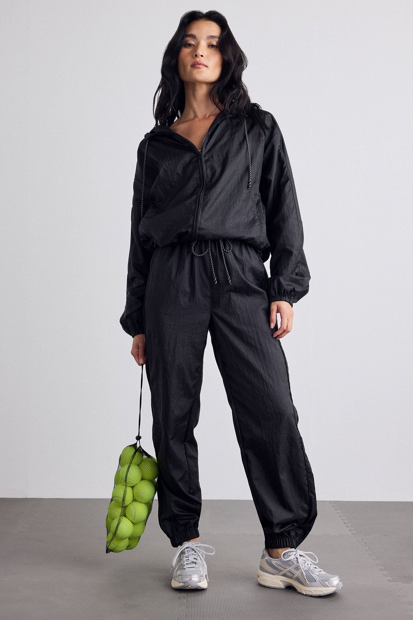 A model stands in a studio wearing a black lightweight zip-front jacket and matching black pants, holding a green bag made of clustered tennis balls. She is wearing silver sneakers against a plain light gray background.