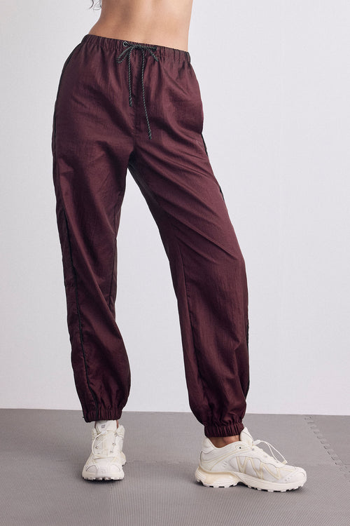 A person wearing dark burgundy jogger-style pants with a drawstring waist and elastic cuffs, paired with white sneakers, shown from the waist down against a light gray studio background.