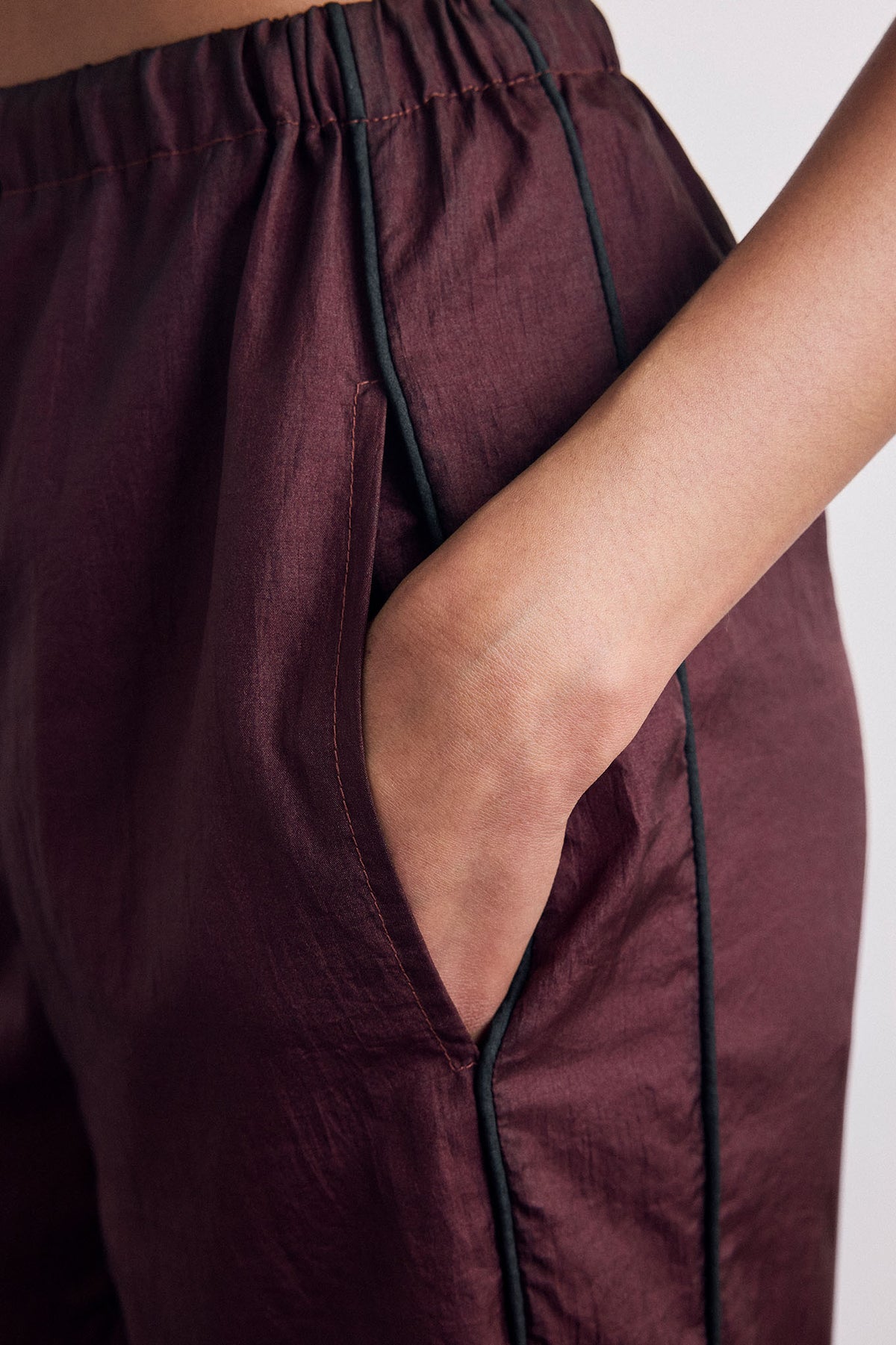 Close-up of a person wearing dark burgundy pants with black piping along the side, showing a hand in a side pocket against a plain light background.