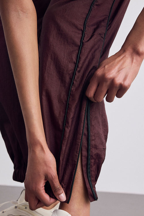Close-up of a person wearing burgundy lightweight fabric with thin dark piping, pulling the side seam slightly with both hands. A white sneaker is partially visible at the bottom.