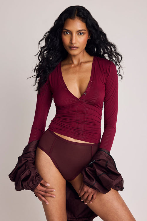 A model wearing a ribbed long-sleeve wrap-style top and matching high-waisted briefs in a deep burgundy color, styled with a burgundy jacket, against a solid light gray studio background.