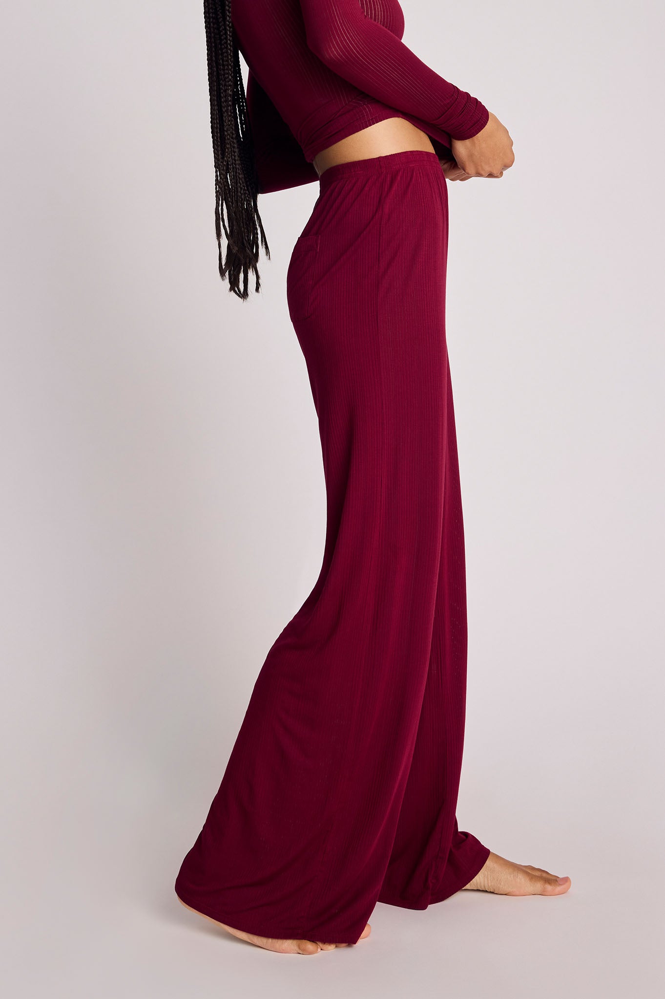 A model wearing a matching deep burgundy two-piece set consisting of a long-sleeve ribbed crop top and high-waisted, wide-leg pleated pants, shown from a side profile against a light gray studio background.