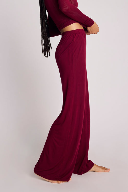 A model wearing a matching deep burgundy two-piece set consisting of a long-sleeve ribbed crop top and high-waisted, wide-leg pleated pants, shown from a side profile against a light gray studio background.