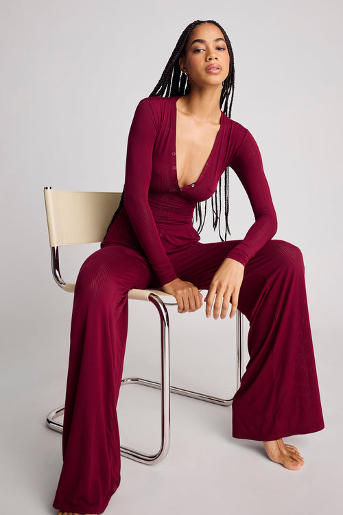 A woman with long black braids sits on a modern chair wearing a deep burgundy long-sleeve top with a plunging V-neckline and pants with wide legs, photographed against a light gray studio background.
