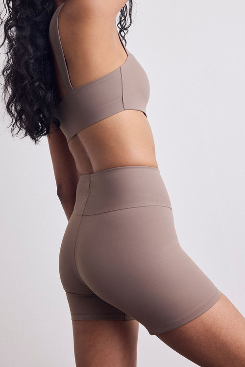 A close-up side view of a model wearing a matching athletic set consisting of a sports bra and high-waisted biker shorts in a solid taupe color, set against a plain light gray background.