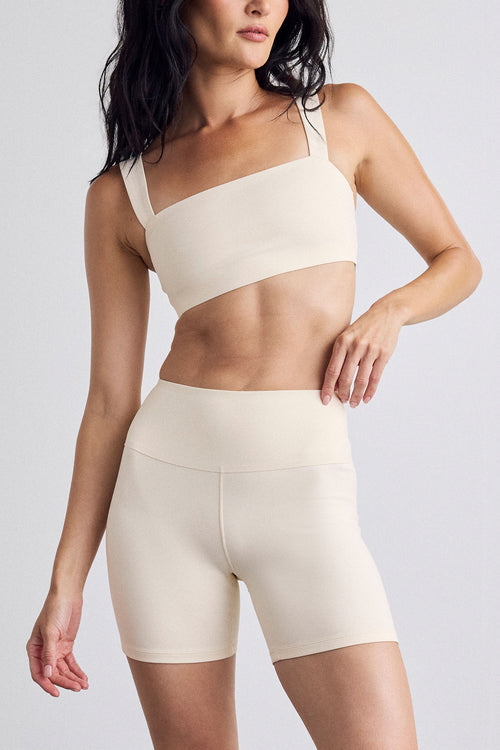 A woman modeling a matching cream activewear set consisting of a sports bra and high-waisted biker shorts against a light gray studio background.