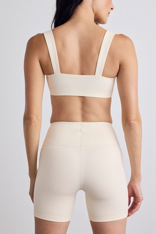Rear view of a model wearing a matching cream sports bra and high-waisted bike shorts against a light gray studio background.