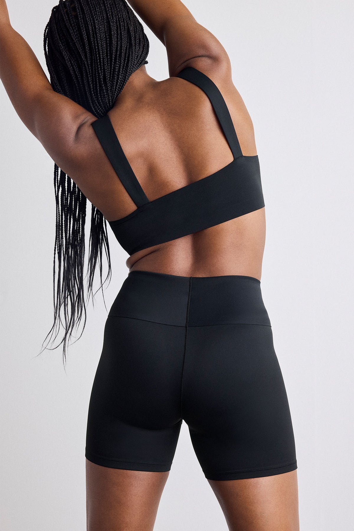 Rear view of a model wearing a black sports bra and matching high-waisted black biker shorts against a light gray studio background.