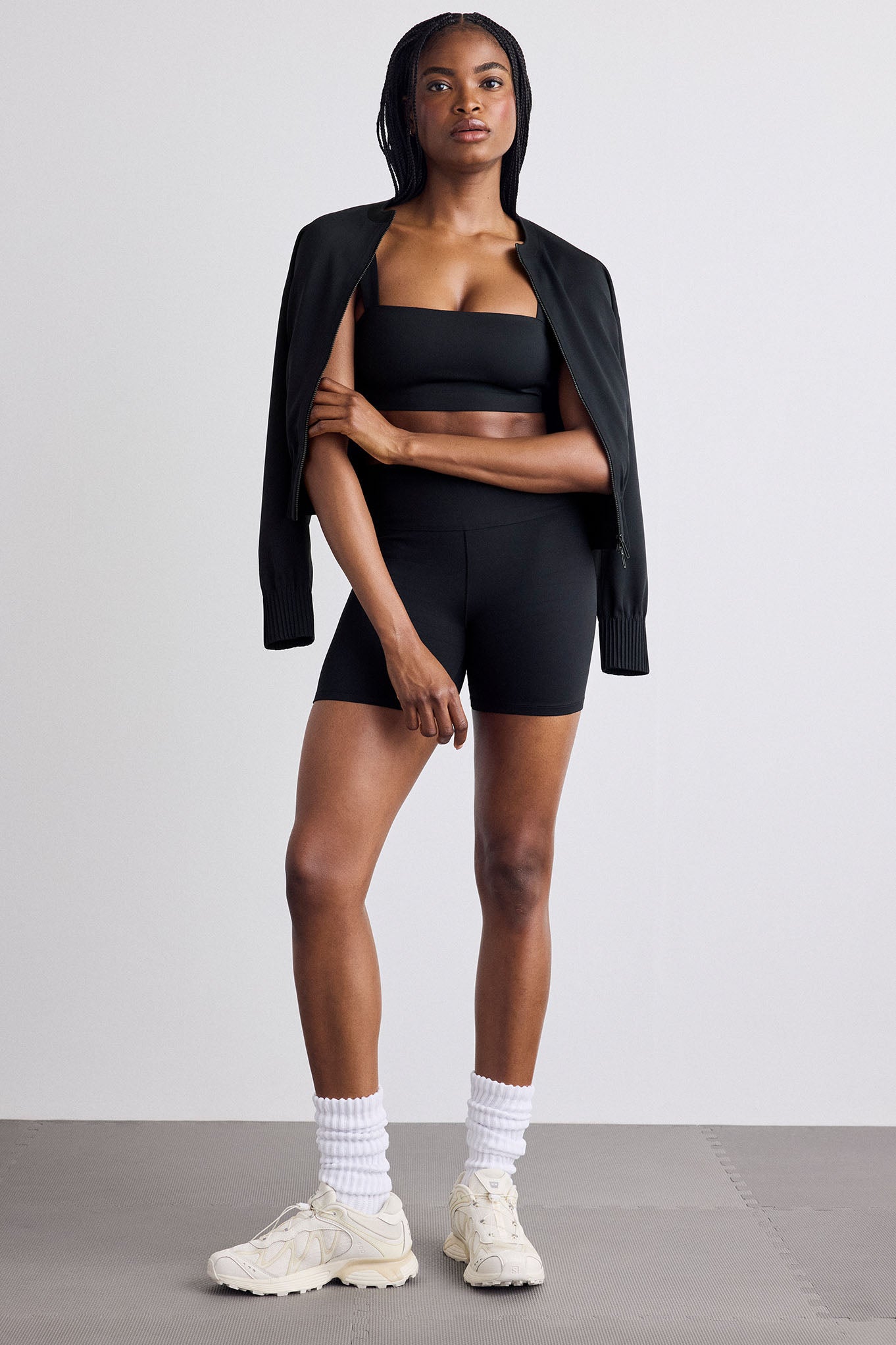 A model stands in a studio wearing a black athletic set consisting of a square neck bra top, high-waisted biker shorts, and an open black bomber draped over her shoulders, paired with white socks and off-white sneakers.