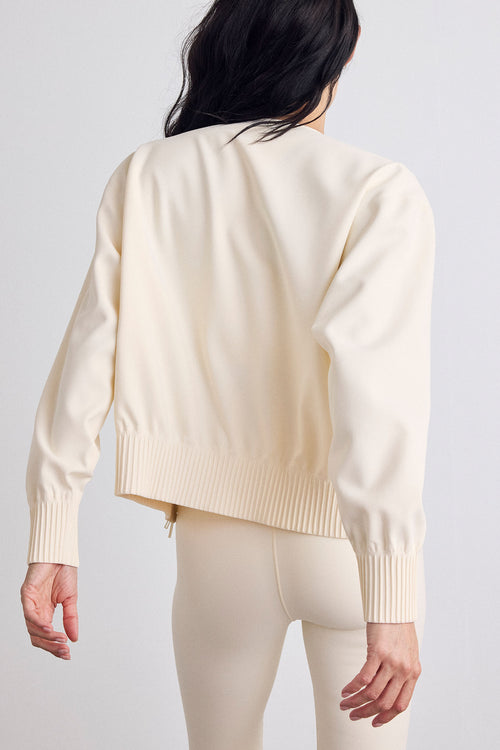 A model shown from the back wearing a cream-colored bomber with ribbed cuffs and hem, paired with matching cream leggings, against a light gray studio background.