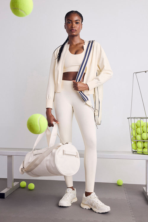 A woman stands in a studio setting wearing a cream-colored athletic set consisting of a crop top, leggings, and zip-up jacket, carrying a striped shoulder bag and holding a large cream duffel bag. Tennis balls are scattered around, with one floating near her head and another in a wire basket on the right.