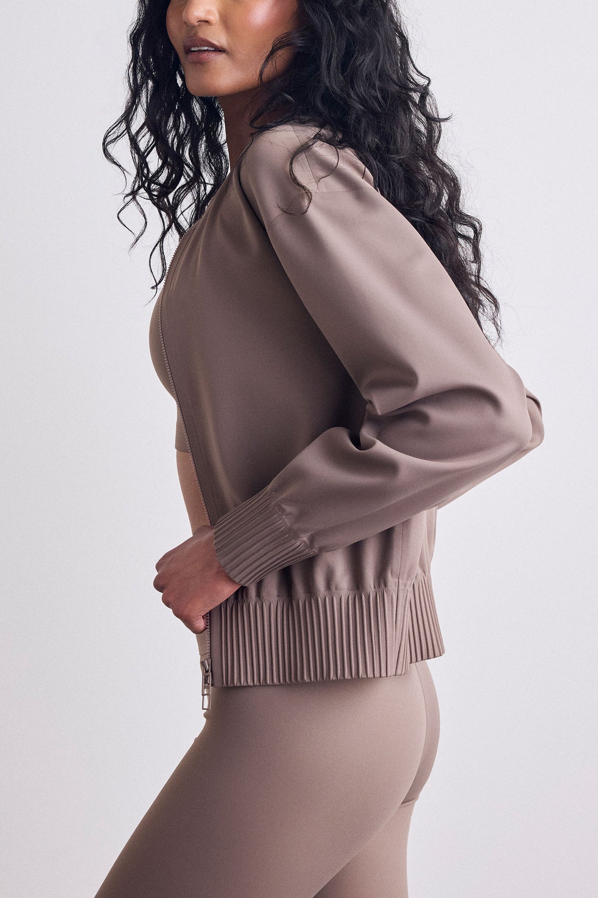 A woman wearing a taupe bomber jacket and matching leggings, shown from a side profile against a light gray studio background.