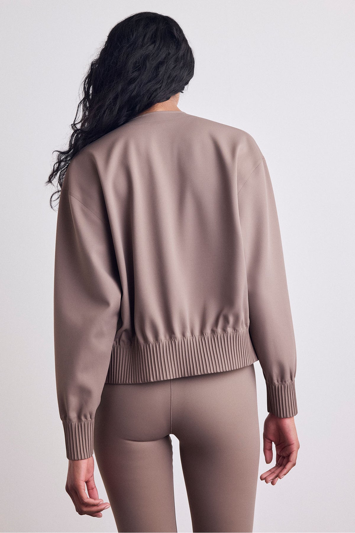 A woman shown from the back wearing a taupe bomber and matching leggings against a light gray studio background.