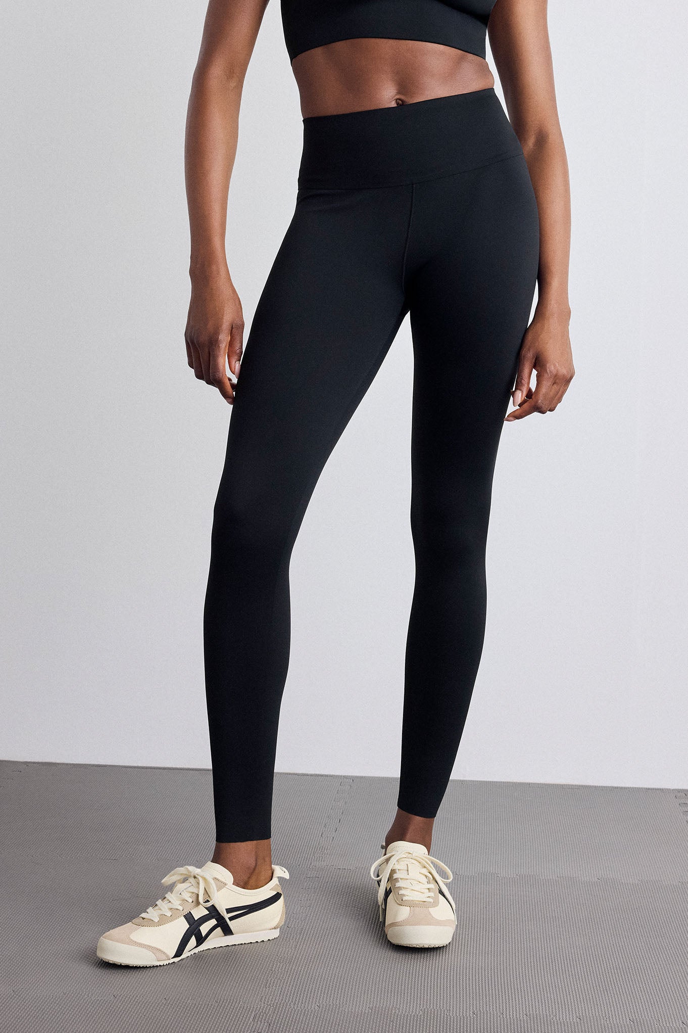 A model wearing black high-waisted athletic leggings and cream sneakers with black stripes, standing against a light gray studio background.