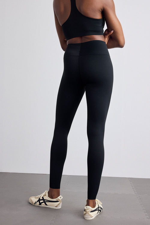 A model from behind wearing black high-waisted leggings, a black sports bra, and beige and black sneakers, standing on a gray floor against a light gray wall.