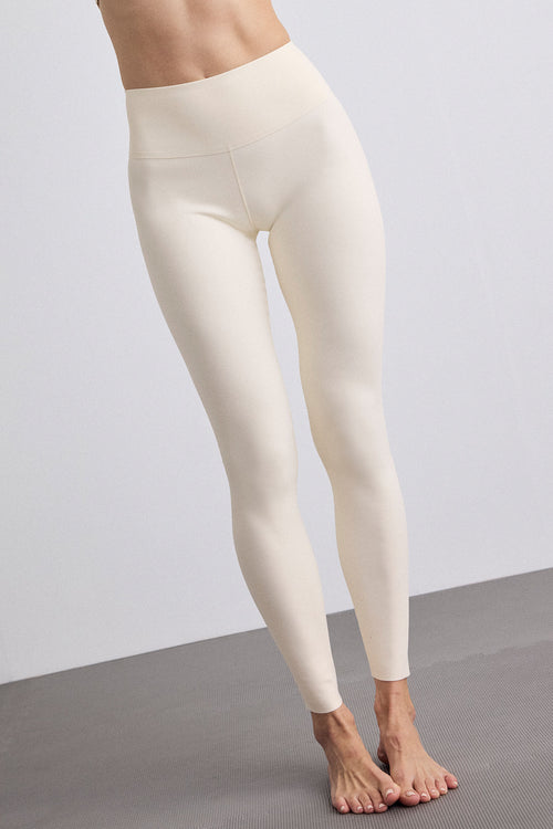 A model wearing high-waisted, full-length leggings in a cream color, standing barefoot on a gray floor against a light gray wall.