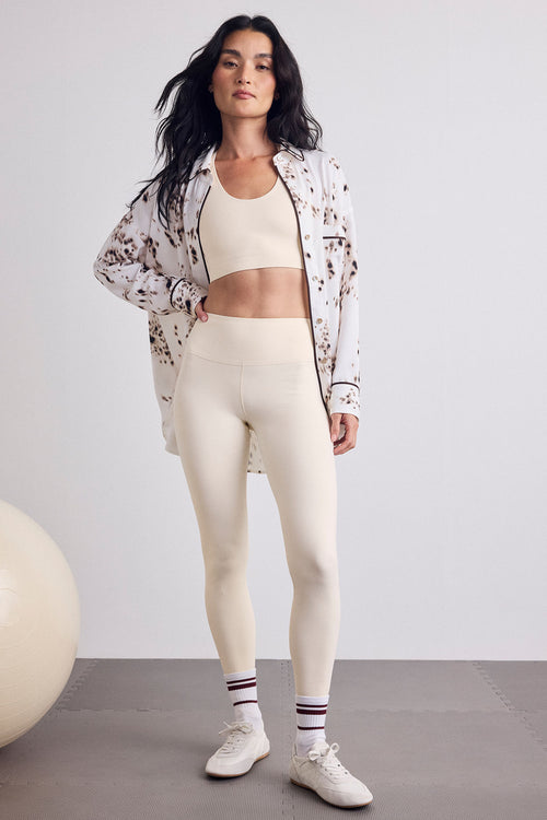A woman modeling a matching activewear set featuring a cream sports bra and high-waisted leggings, styled with a white and brown shirt, white socks, and white sneakers.