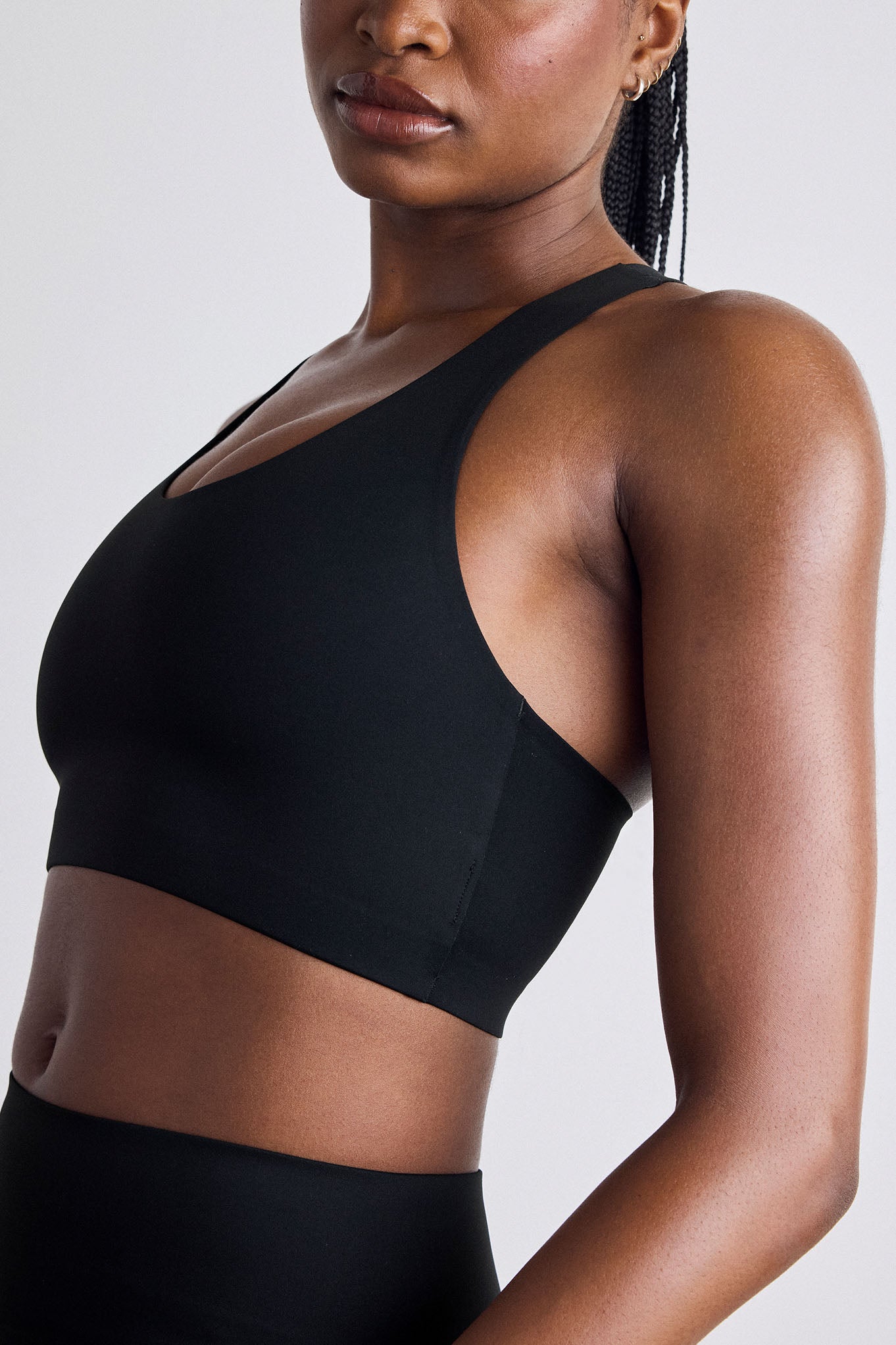 A close-up of a woman wearing a black sports bra and matching black bottoms against a light gray studio background.