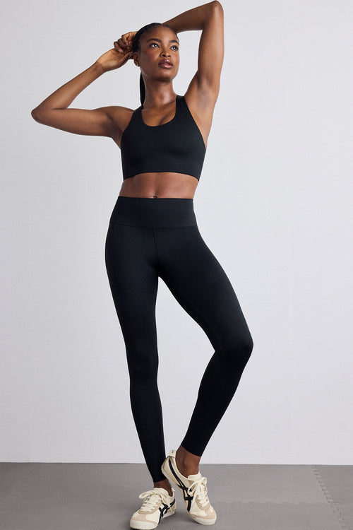 A woman modeling a matching black athletic set consisting of a sports bra and high-waisted leggings, paired with white sneakers, against a light gray studio background.