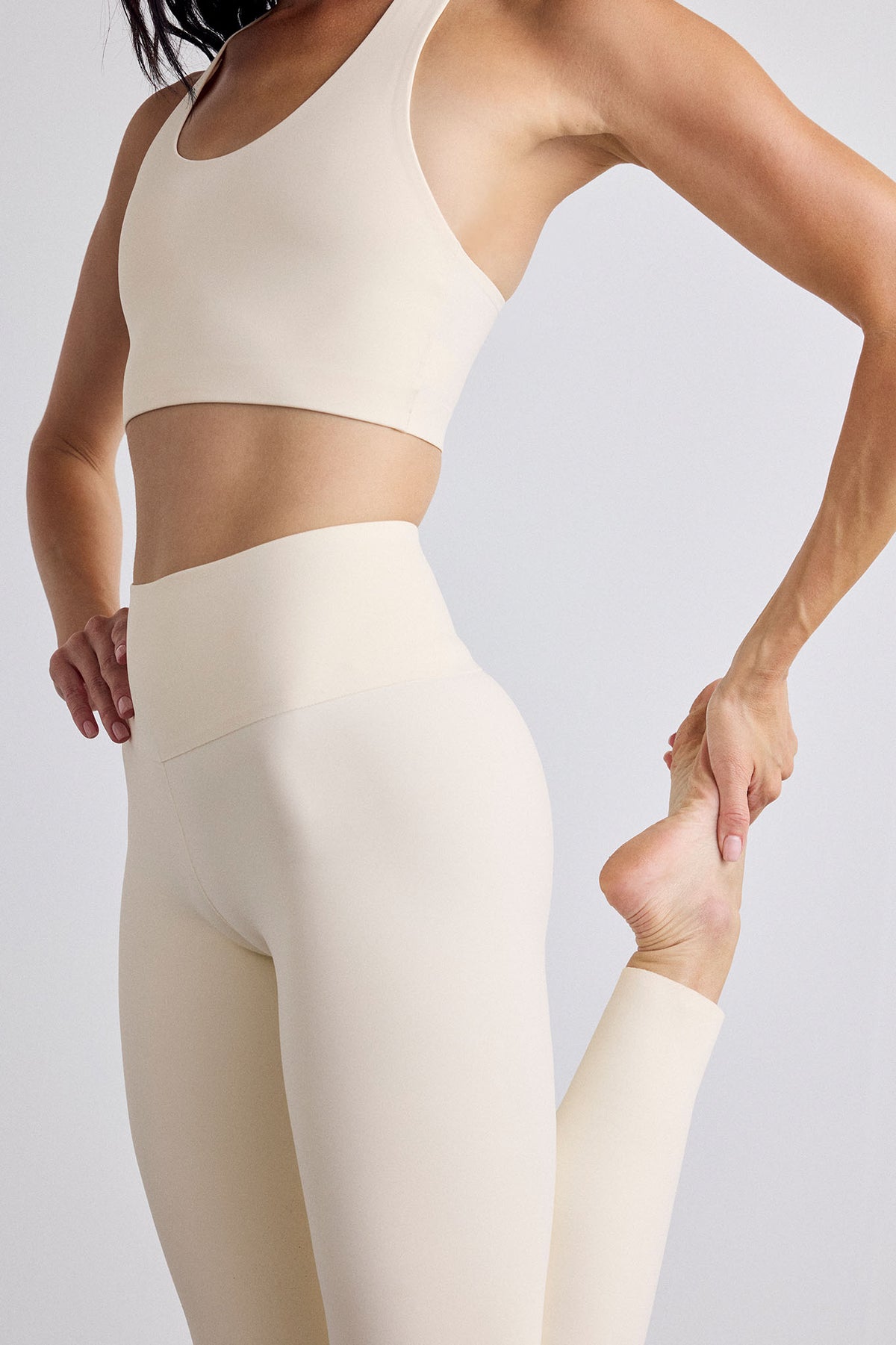 A woman wearing a matching cream-colored activewear set, featuring a sports bra and high-waisted leggings, stretching her leg against a light gray studio background.