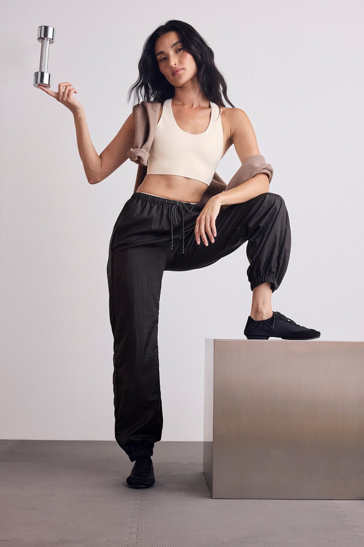 A woman poses in a studio wearing a beige sports bra, black jogger pants, and black sneakers, holding a silver dumbbell.