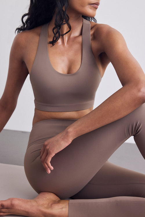 A woman wearing a matching taupe-brown athletic set consisting of a scoop-neck sports bra and high-waisted leggings, posing in a minimalist studio setting.
