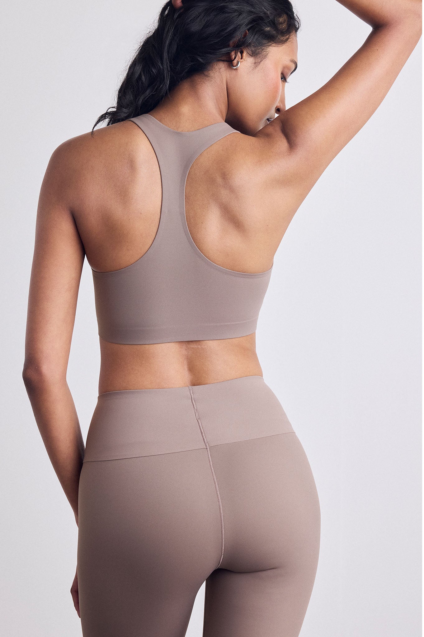 Rear view of a woman wearing a matching taupe-brown sports bra and high-waisted leggings set against a light gray studio background.