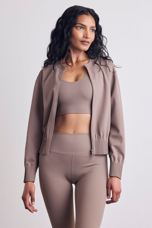 A woman modeling a matching taupe activewear set consisting of a cropped sports bra, ribbed zip-up bomber jacket, and high-waisted leggings against a light gray studio background.