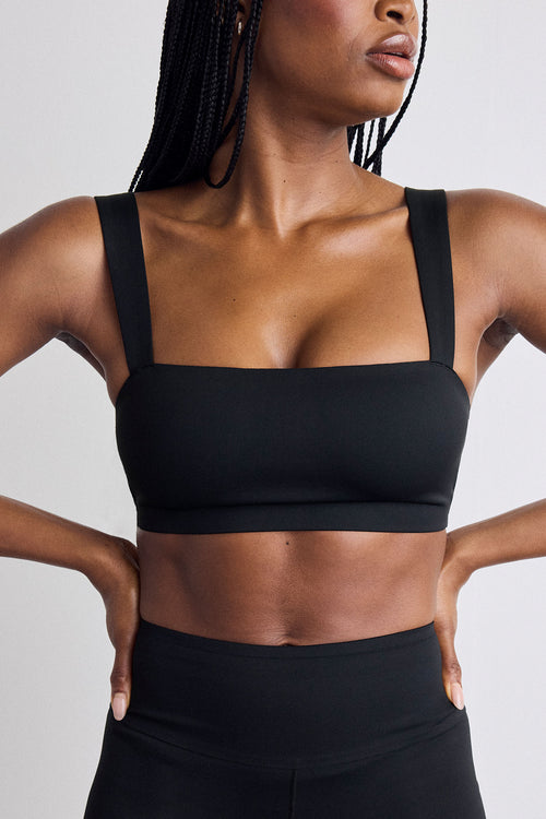 A close-up of a model wearing a black sports bra with wide shoulder straps and a square neckline, paired with black high-waisted leggings, against a light gray studio background.
