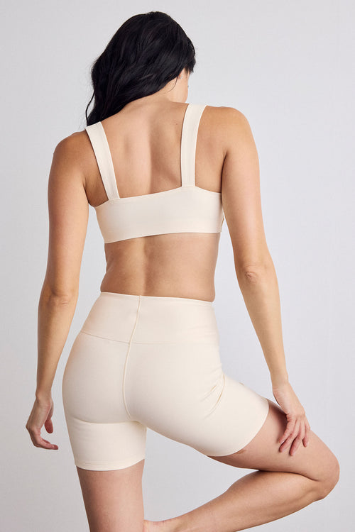 A woman shown from the back wearing a matching cream sports bra and high-waisted biker shorts set against a light gray studio background.