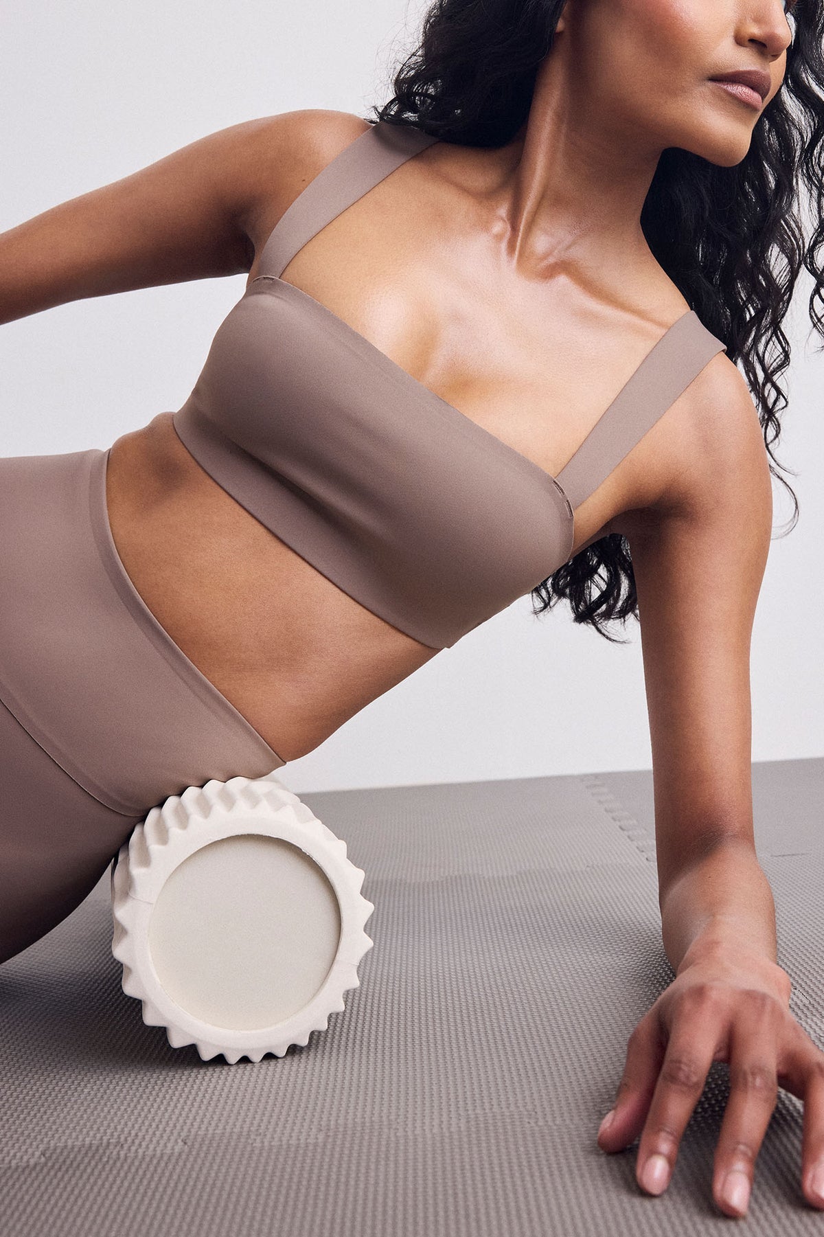 A woman in a taupe sports bra and matching leggings poses on a gray yoga mat, using a white textured foam roller on her lower back. 