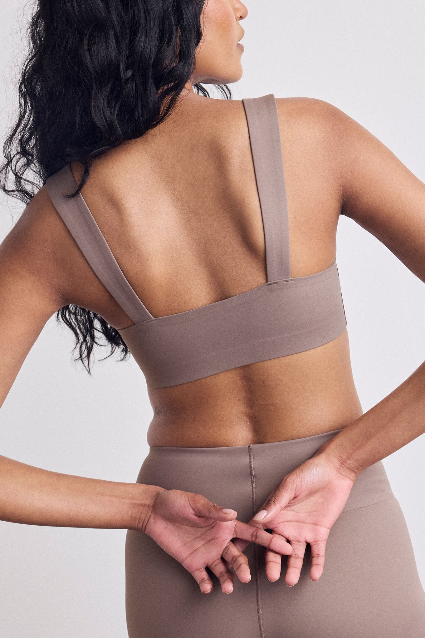 Rear view of a woman wearing a taupe-brown sports bra and matching leggings against a light gray studio background.
