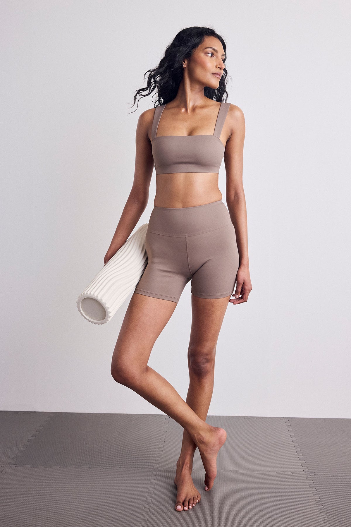 A woman modeling a matching taupe activewear set consisting of a sports bra and high-waisted shorts, holding a white textured foam roller against a neutral studio backdrop.