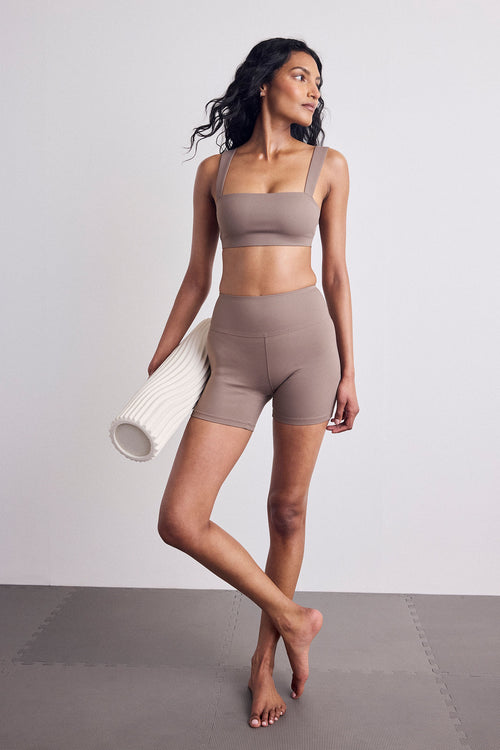 A woman modeling a matching taupe activewear set consisting of a sports bra and high-waisted shorts, holding a white textured foam roller against a neutral studio backdrop.